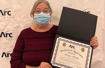Arc/Morris Residential Manager Susan Many stands in front of a white banner with the Arc/Morris logo holding her Certificate of Appreciation for 15 years of service.