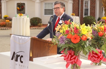 Senator Anthony Bucco speaks at a podium outside in front of the F.M. Kirby Administrative Center; there are two bouquets of flowers in glass vases adorned with red bows on the table in the foreground.