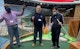 Three men are standing in front of a mini golf hole at an indoor mini golf location. One man is raising his arm and putter in the air.