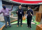 Three men are standing in front of a mini golf hole at an indoor mini golf location. One man is raising his arm and putter in the air.
