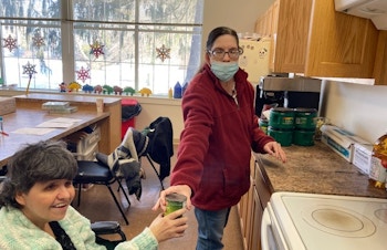 A woman in a wheelchair hands another woman a can to put in a kitchen cabinet.