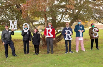 Seven individuals we serve stand in front of a tree holding large letters which spell out Morris.