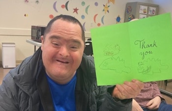 A man smiles as he holds up a green thank you card he made.