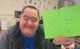 A man smiles as he holds up a green thank you card he made.