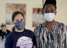 Two females standing next to one another wearing protective masks.